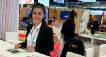 Hôtesse d’accueil souriante derrière un comptoir d’information du salon Mountain Planet, prête à renseigner les visiteurs.