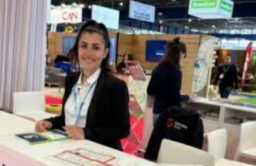 Hôtesse d’accueil souriante derrière un comptoir d’information du salon Mountain Planet, prête à renseigner les visiteurs.