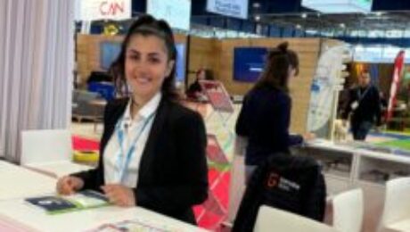 Hôtesse d’accueil souriante derrière un comptoir d’information du salon Mountain Planet, prête à renseigner les visiteurs.