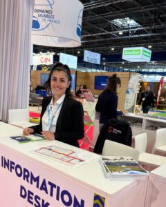 Hôtesse d’accueil souriante derrière un comptoir d’information du salon Mountain Planet, prête à renseigner les visiteurs.