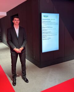 Hôte d’accueil en costume noir, debout à l’entrée d’un événement professionnel avec un écran affichant le programme du salon Mountain Planet.